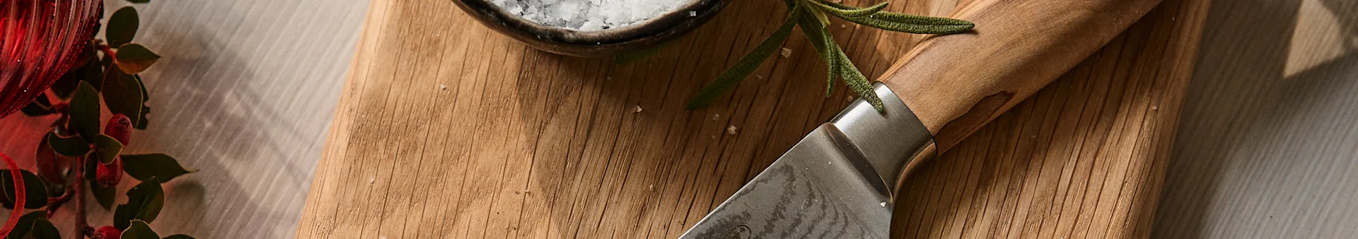 Kniv, rosmarin og havsalt på et skjærebrett i tre ved siden av en rød festlig dekorasjon.