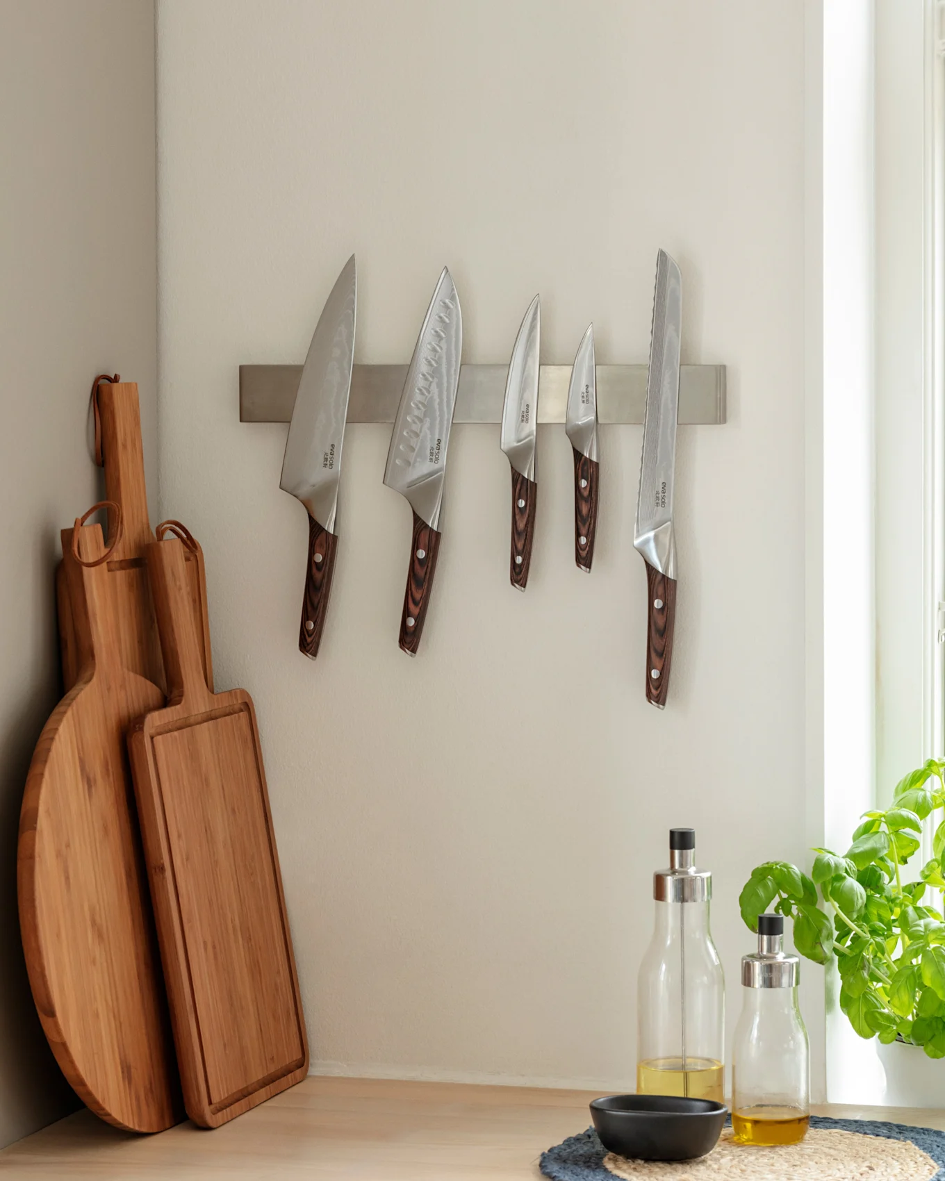 Kjøkkenhjørne med fem kniver på en magnetlist, to skjærebrett i tre og to flasker olivenolje på en trekjøkkenbenk ved siden av en basilikumplante.