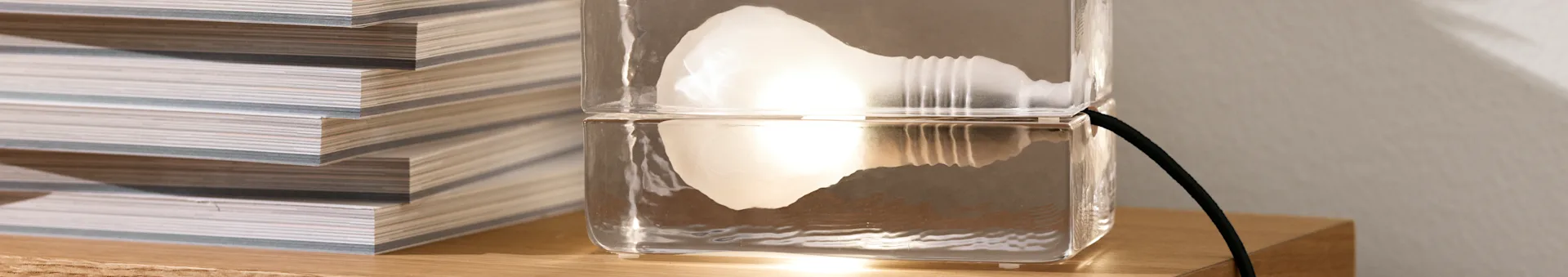 En unik klar lampe, laget av to stablede rektangulære blokker, med et glødende internt lys, ved siden av en stabel magasiner på en treoverflate.