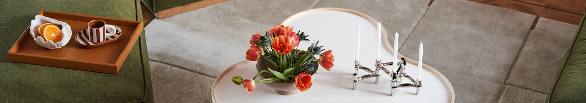 Grønn sofa, salongbord med oransje tulipaner og sølv lysestake. Brett på sofaen har appelsinskiver og stripet kopp.
