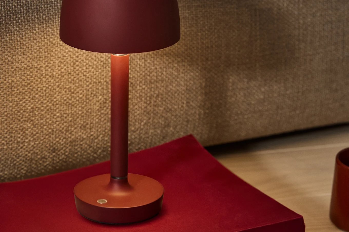 En dyprød minimalistisk bordlampe med et varmt skjær, plassert på en rød overflate mot en teksturert brun bakgrunn.