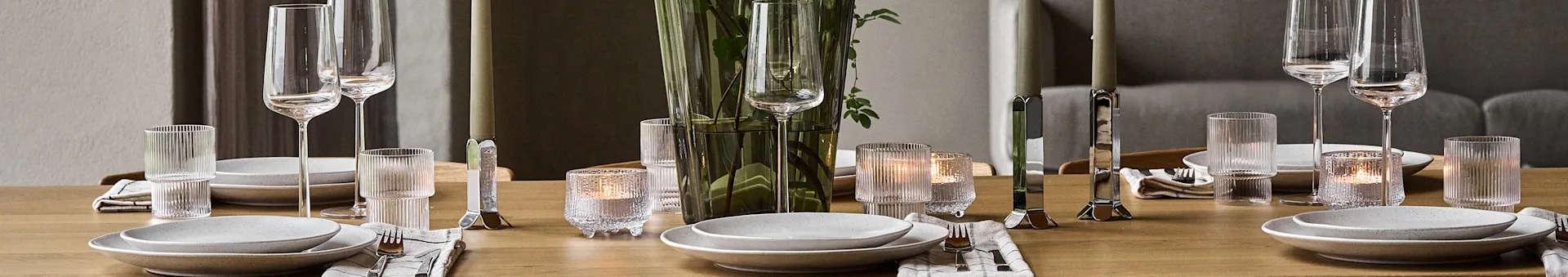 Et spisebord i tre dekket med hvite tallerkener, sølvbestikk, klare glass, tente lys og en grønn vase med blader.