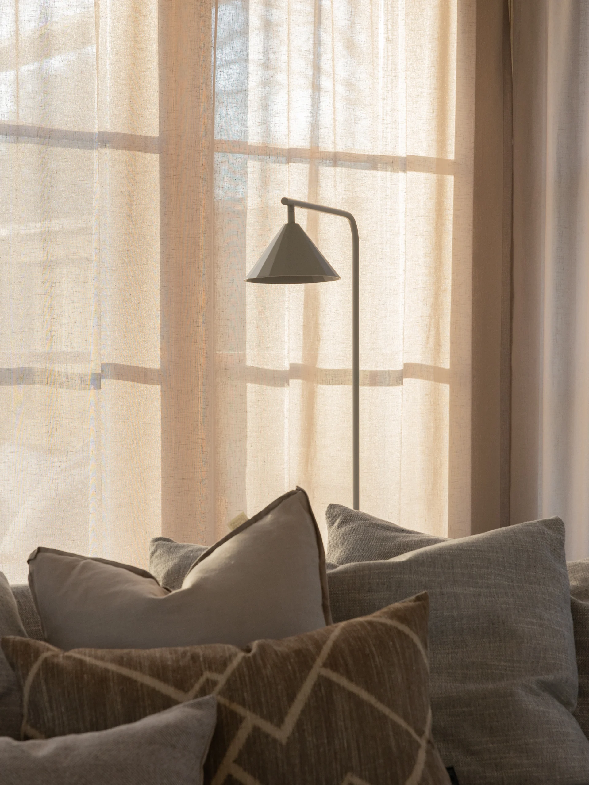 En moderne grå gulvlampe foran solbelyste gardiner, med ulike puter på en sofa i forgrunnen.