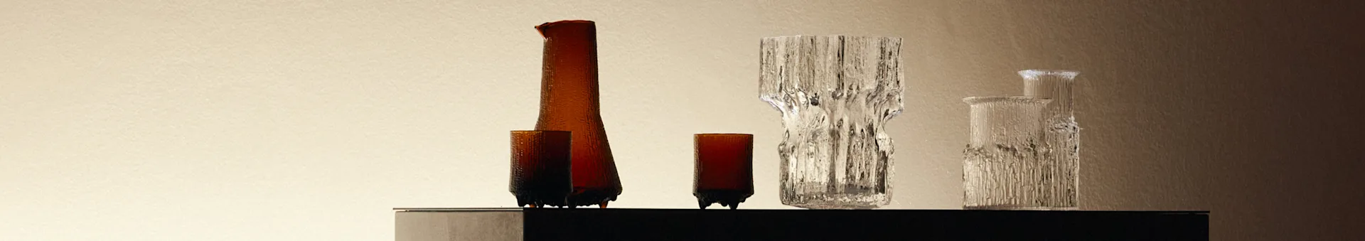 En samling teksturerte glassvaser og flasker, ravfargede og klare, på en mørk overflate mot en lys bakgrunn.