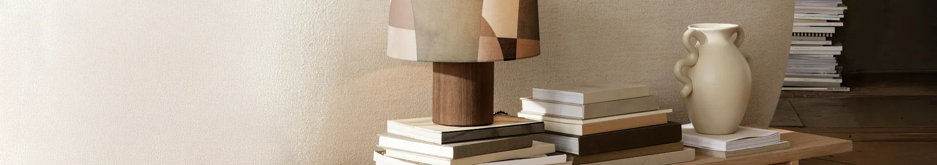 En høy lampe med mønstret stoffskjerm og en lys beige vase står på et trebord, omgitt av stabler med bøker mot en teksturert vegg.