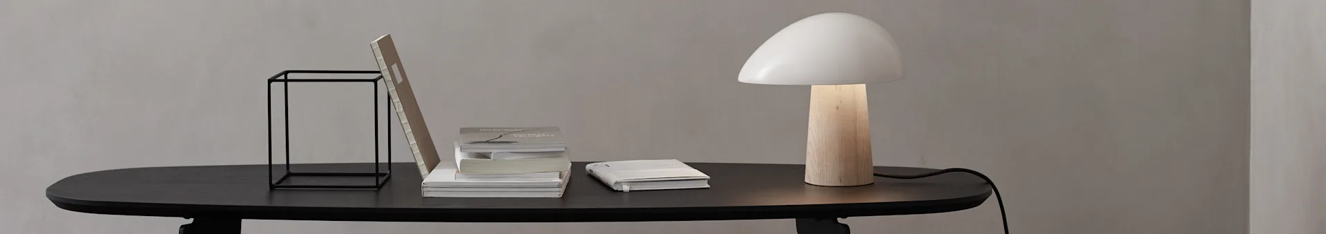 Et minimalistisk skrivebord med et svart bord, svart metallstativ, stablede bøker og en lampe med trebunn og hvit soppskjerm.