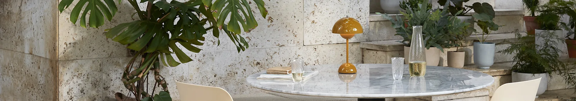 En sennepsgul bordlampe og klar glassvase på et marmorbord med en monstera-plante til venstre og potteplanter på trappen til høyre.