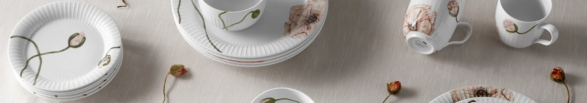 Hvite riflede porselenstallerkener, skåler og kopper med valmueblomstdesign på lyst stoff, med tørkede valmuefrø.
