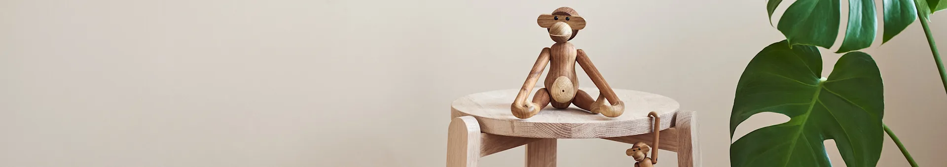 En stor treape-figur sitter på lys trekrakk, en mindre henger under, ved siden av en grønn plante.
