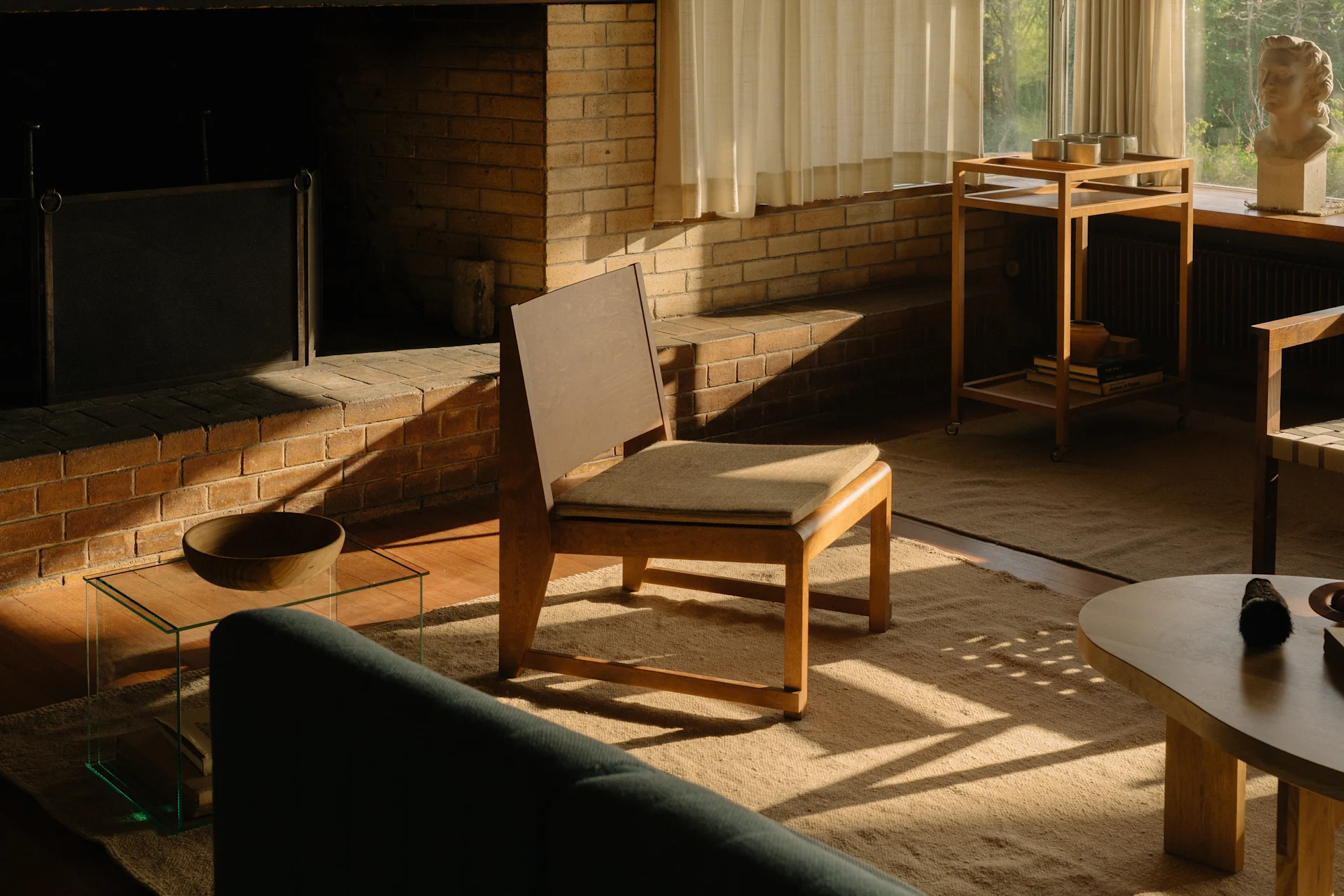 En solfylt stue med en mursteinspeis, en trestol og en grønn sofa, med en bysteskulptur ved vinduet.