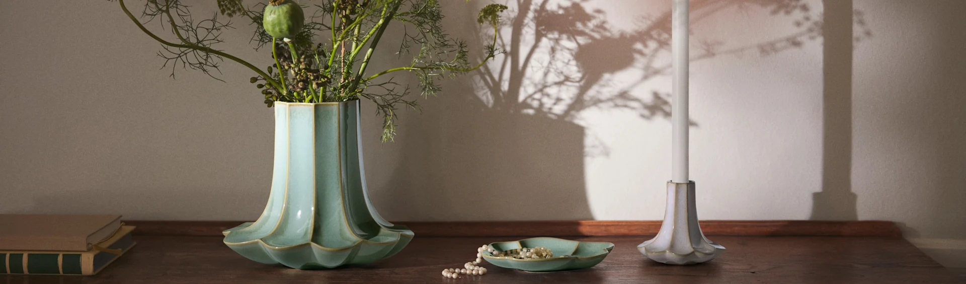 En riflet turkis vase med løv, et perlekjede i en matchende skål, et hvitt stearinlys i en riflet holder, og bøker på en treoverflate.