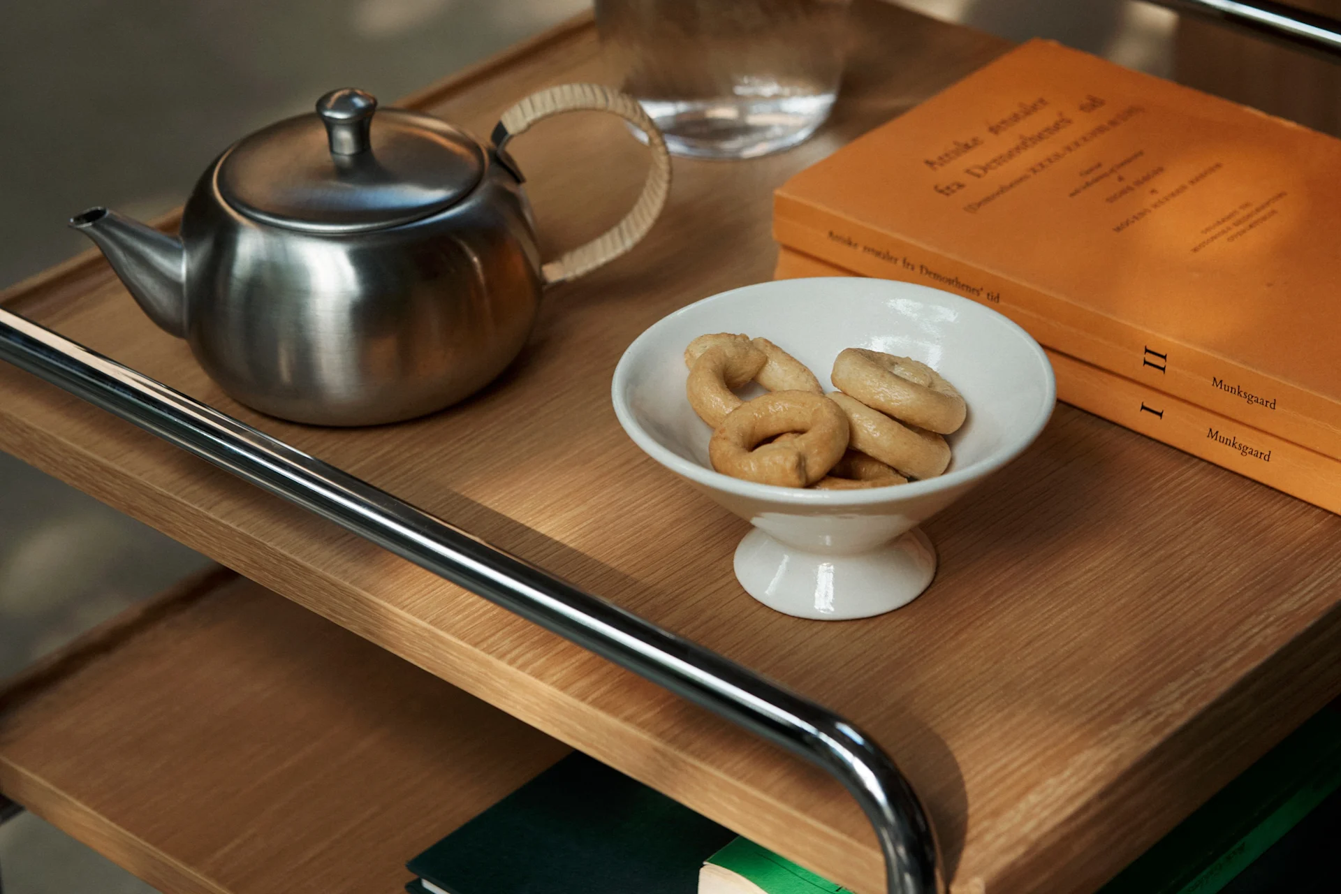 En tekanne i rustfritt stål, en hvit bolle med ringkjeks og to oransje bøker på en trehylle.