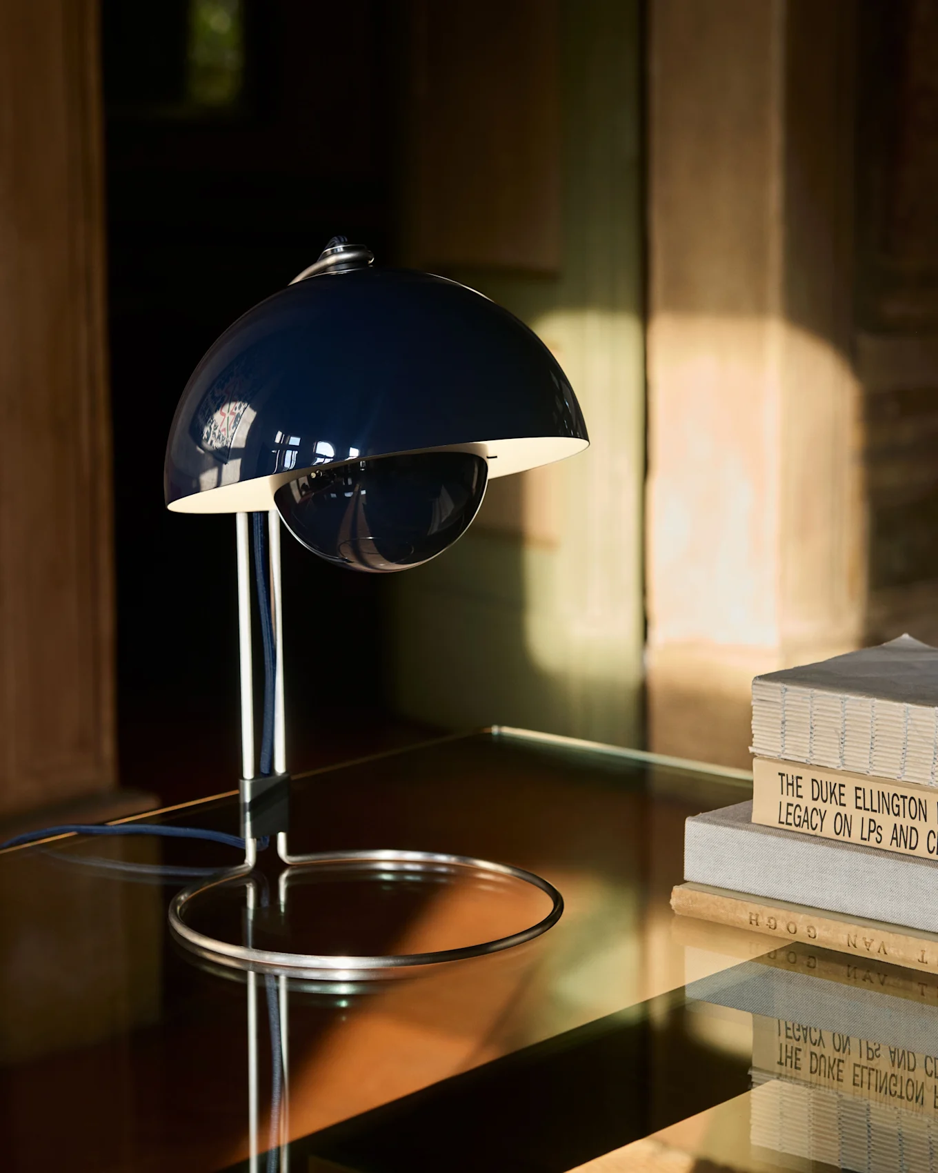 En mørkeblå, klokkeformet bordlampe med krombase står på et reflekterende bord ved siden av en stabel bøker.