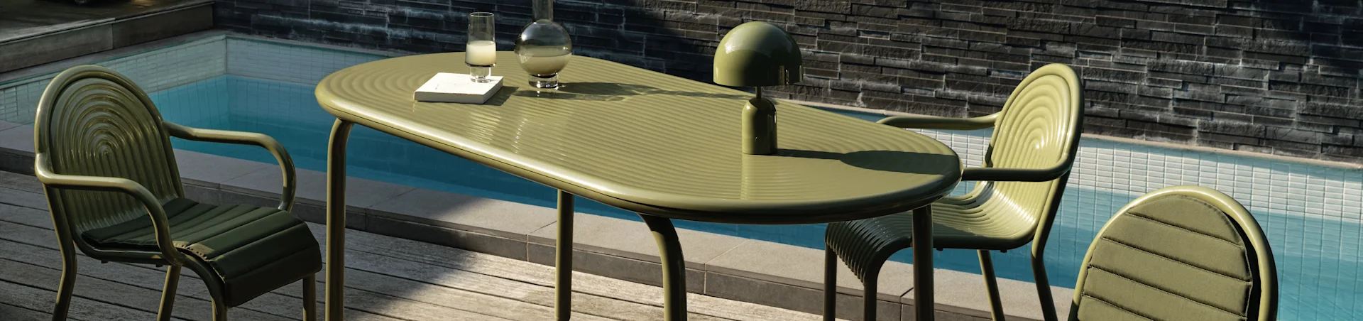 Olivengrønn utendørs spisesett ved et basseng med tre stoler, et ovalt bord, en bok, en drink, en karaffel og en lampe.