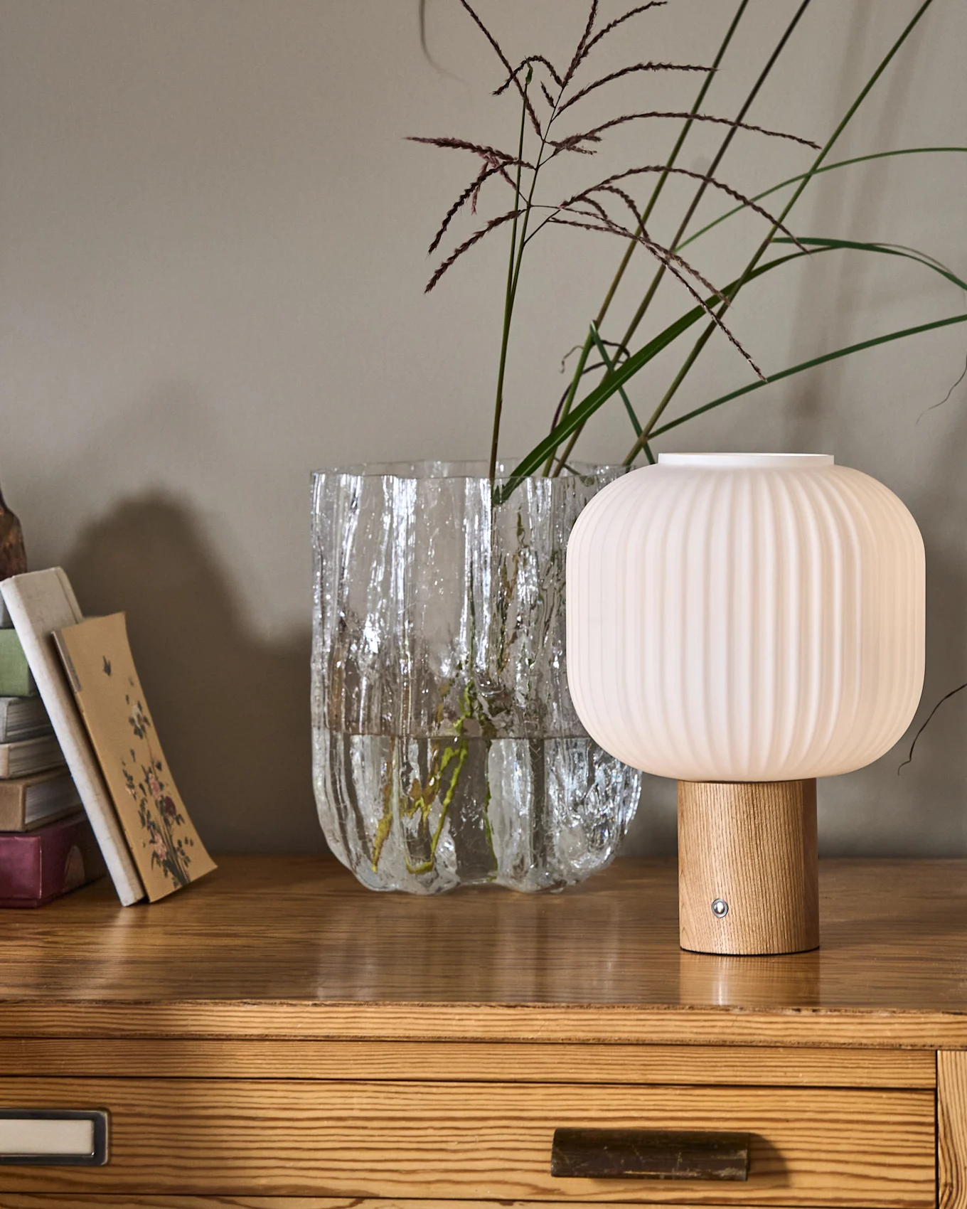 En ribbet, soppformet lampe med trefot ved siden av en teksturert glassvase med planter og en stabel bøker på en kommode av tre.