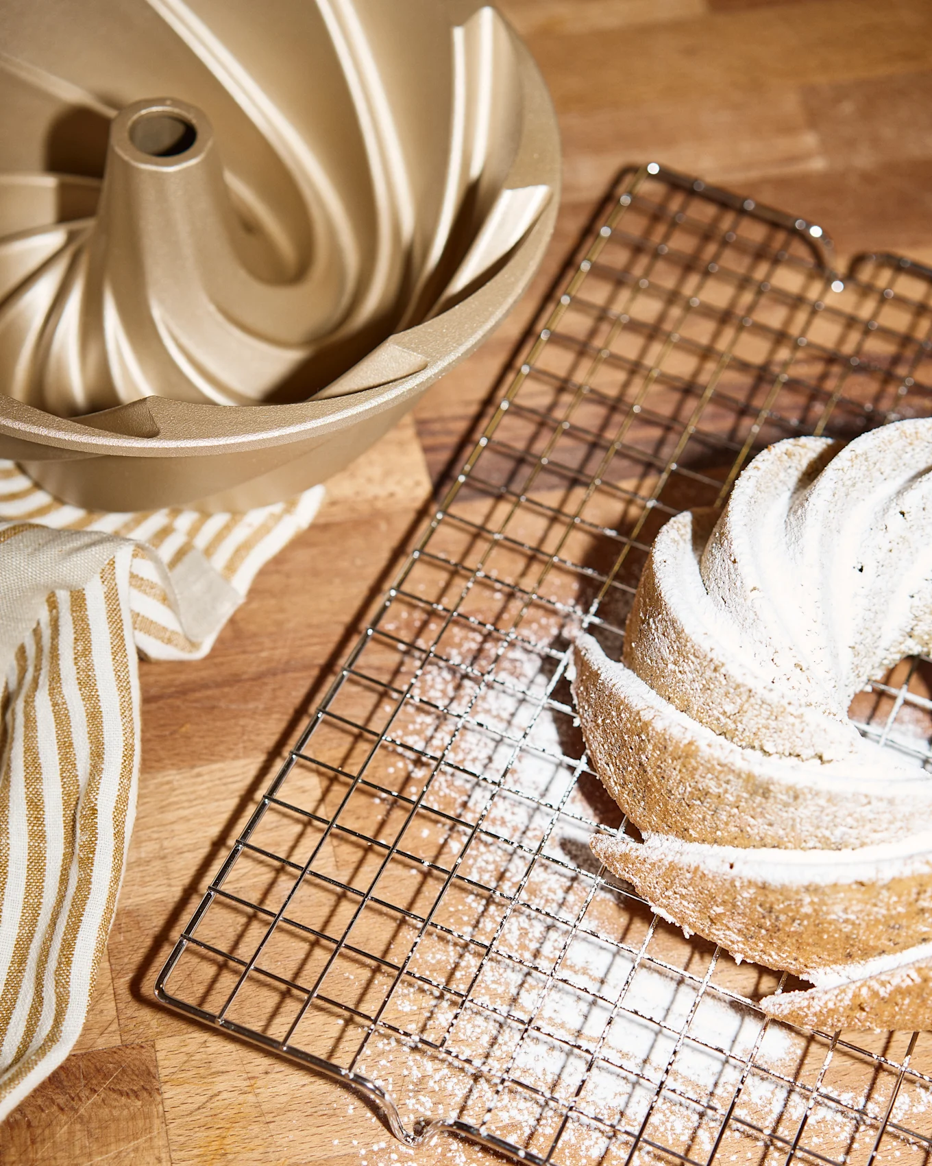 En gyllen bundt kakeform, en avkjølingsrist med en pudderdryss-dekket bundtkake og et stripete håndkle på en treoverflate.