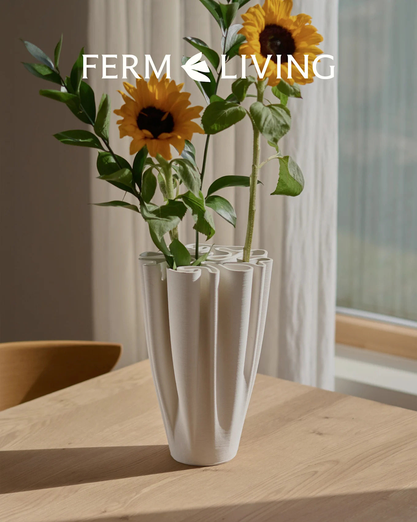 Solsikker i en hvit, ribbet bølgeformet vase på et trebord. Ferm Living logo overlay.