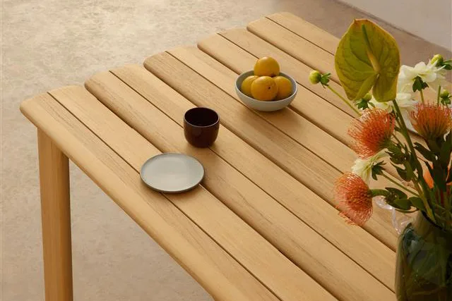 Lyst trelamellbord med en bolle sitroner, mørkt krus, liten tallerken og en vase med oransje og hvite blomster.