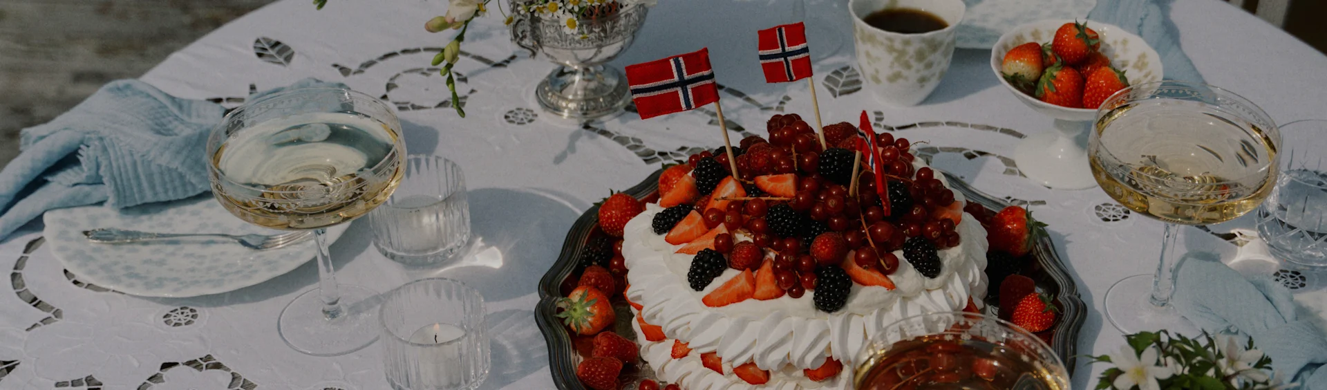 Festlig bord med pavlovakake toppet med bær og norske flagg, champagne, jordbær og kaffe.