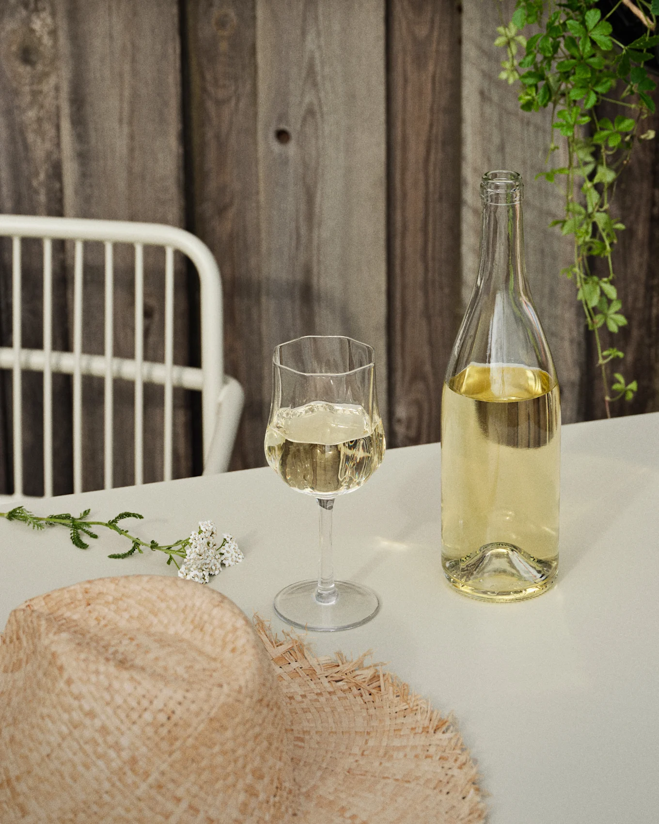 En flaske og et glass hvitvin på et bord med en stråhatt og hvite blomster, mot en trevegg og grønne slyngplanter.