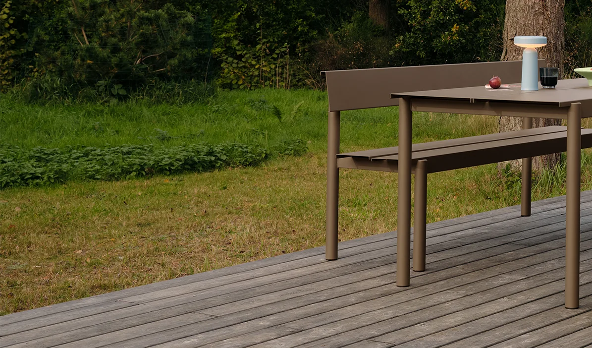 Moderne utendørs spisebord og benk i taupe på en treterrasse, med en bærbar lampe og epler på bordet.