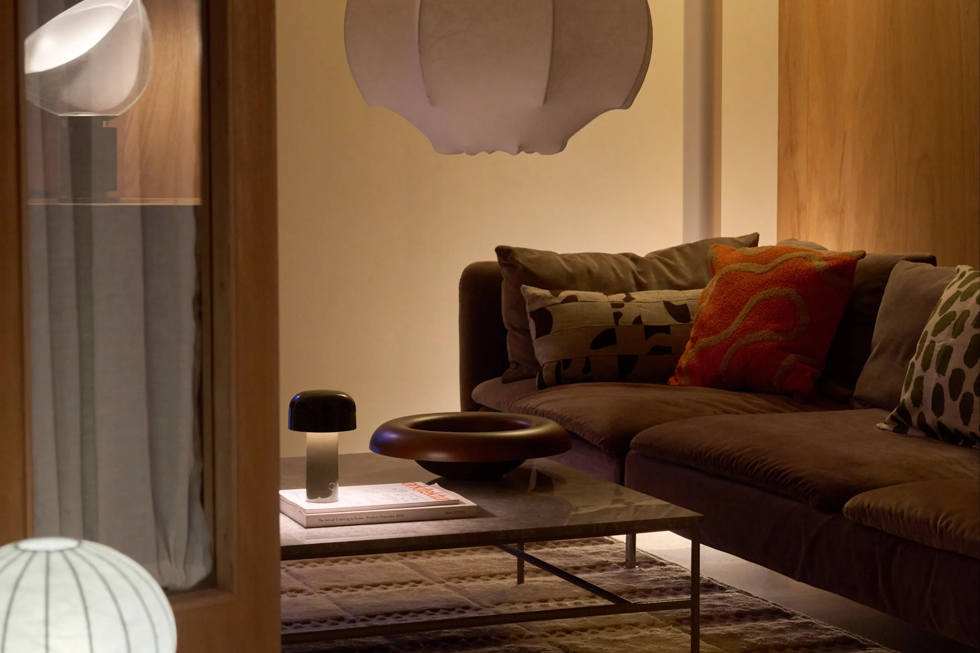 Koselig stue med brun sofa, hvit pendellampe, gulvlampe, salongbord, soppformet lampe og dekorativ bolle.
