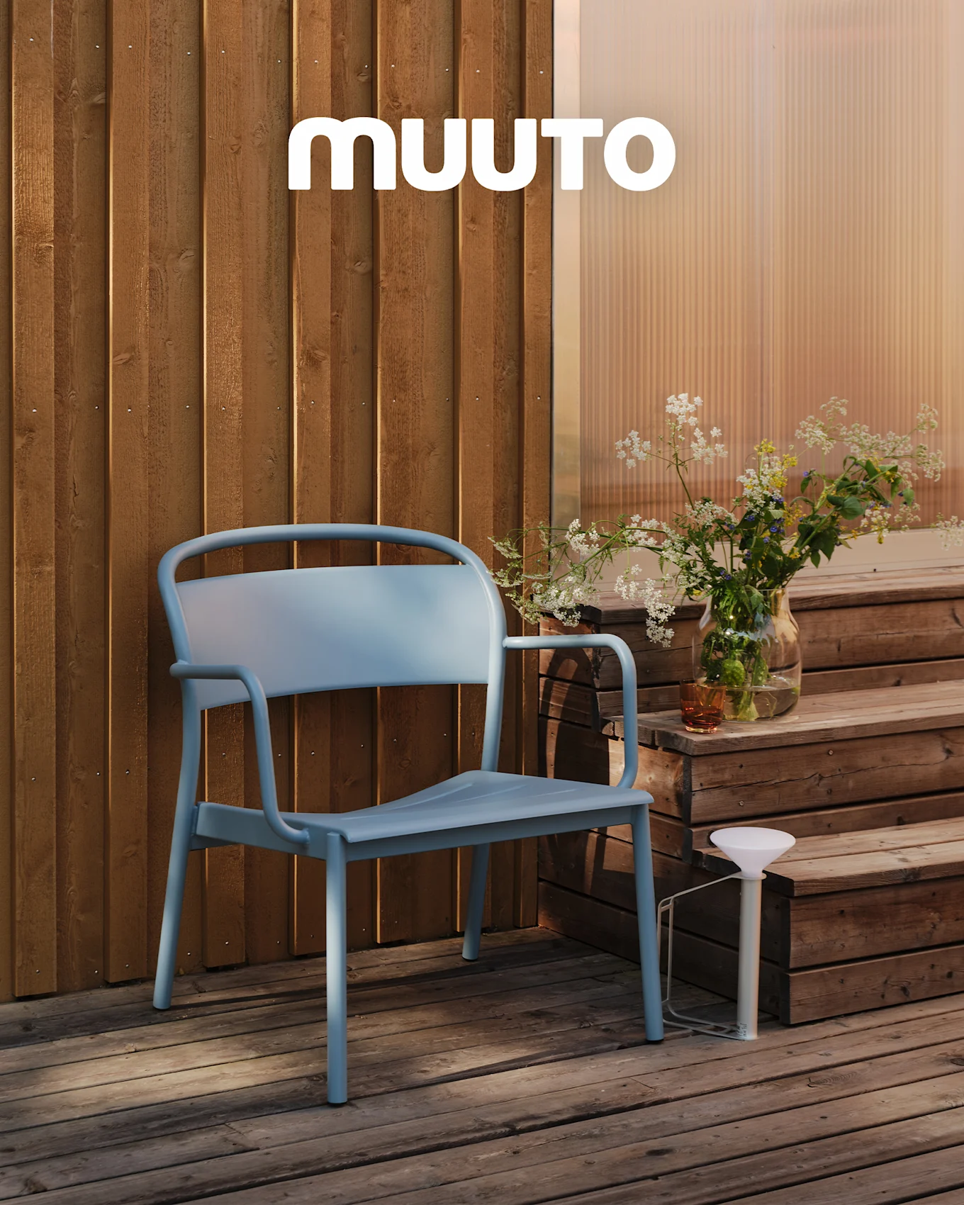 En lyseblå utestol på et tredekk ved trinn med en vase markblomster og en hvit bærbar lampe. MUUTO-logo.
