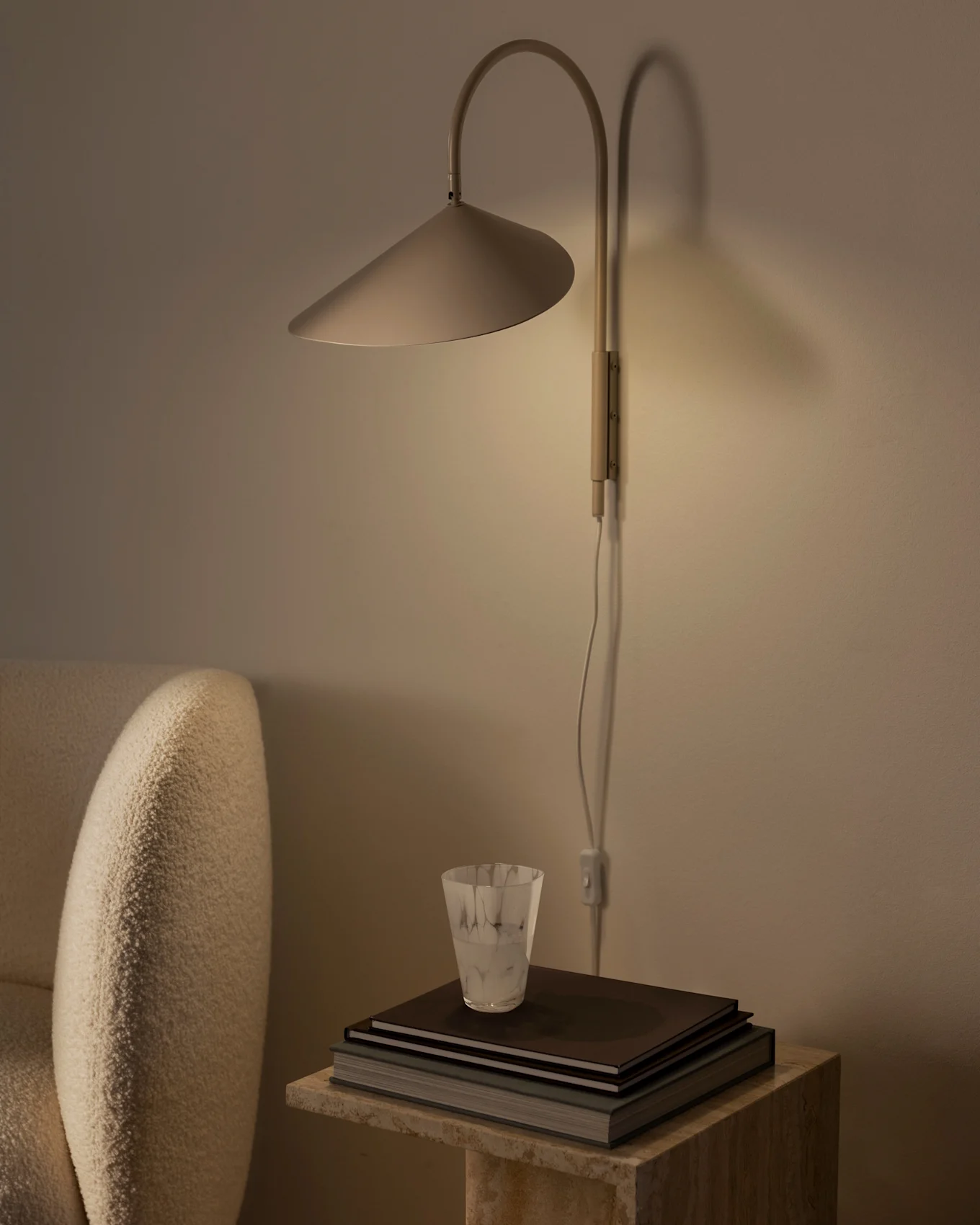 Varmt lysende vegglampe med konisk skjerm over en stabel bøker og et teksturert glass på et steinbord, ved siden av en beige lenestol.