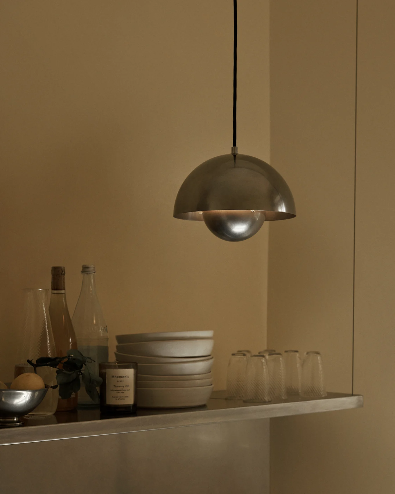 En pendellampe med børstet metalldom henger over en hylle med boller, flasker og glass.