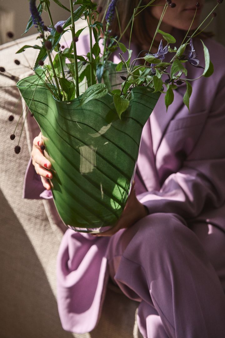 En person iført lilla klær holder den grønne glassvasen Tiggy fra Byon, en vase med plissert, klokkeform i ertegrønt med striper.